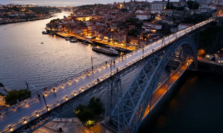 A bridge in Porto, Portugal in the evening with illuminated buildings behind it.
